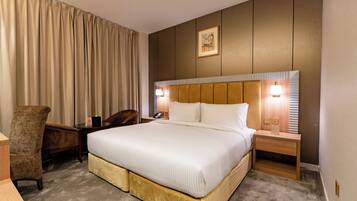 A hotel room with a large bed, a chair, a desk, and a nightstand with a lamp.