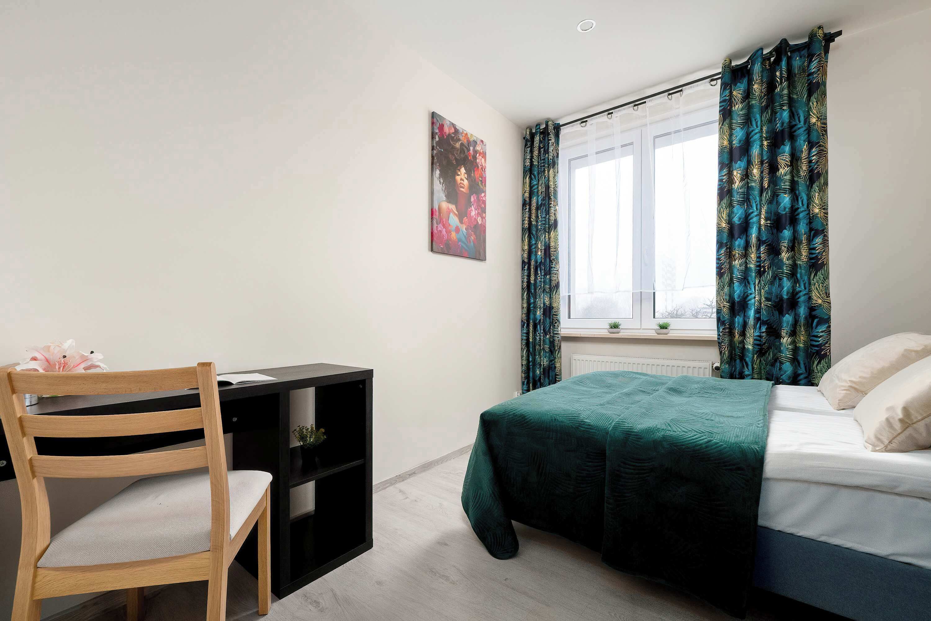 Ein Schlafzimmer mit Bett, Schreibtisch, Stuhl, einem Fenster mit Vorhängen und einem Wandbild.