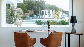 Ein modernes Zimmer mit großem Fenster mit Blick auf einen Pool und Sitzgelegenheiten im Freien. Ein Tisch mit zwei Stühlen, eine Weinflasche und ein Buch sind vorhanden.