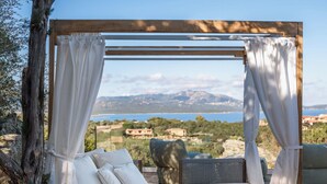 Eine Holzpergola mit weißen Vorhängen, ein Bett mit weißen Kissen und Blick auf eine Küstenlandschaft.