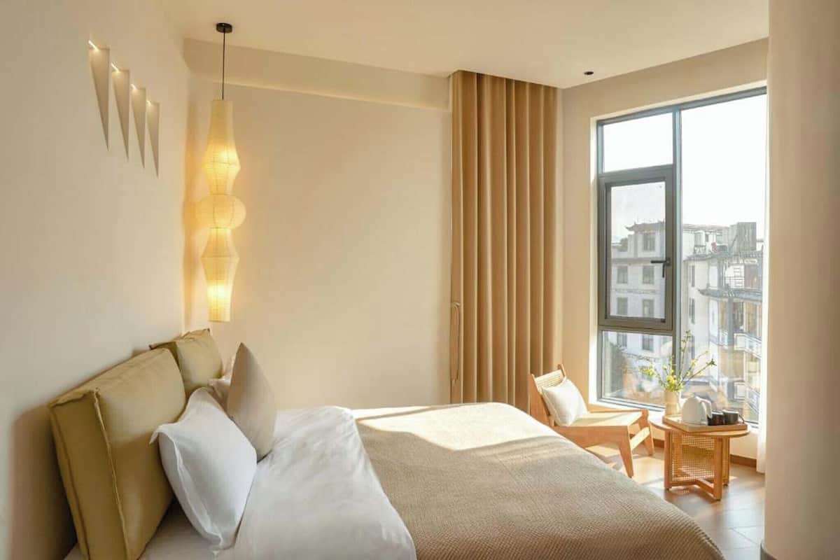 Ein modernes Hotelzimmer mit einem großen Bett, einem Nachttisch mit Lampe sowie Blick auf die Stadt durch ein großes Fenster und einer im Fenster erkennbaren Stadtansicht.