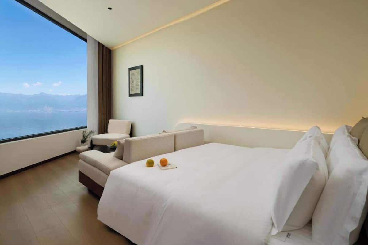 Ein modernes Hotelzimmer mit einem großen Bett, einer Couch und einem Sessel. Ein Fenster bietet Blick auf das Meer und die Berge.
