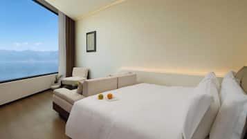 Habitación de hotel moderna con una cama grande, un sofá y una silla. Se ve una ventana con vistas al mar y a las montañas.