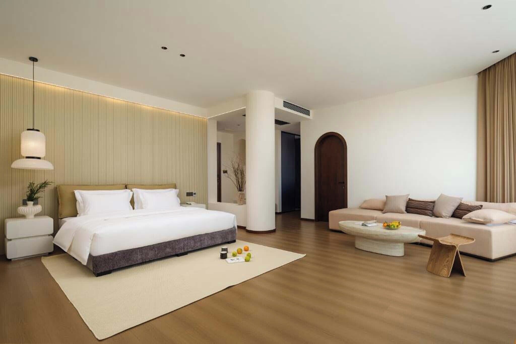 A modern hotel room with a large bed, a sofa, a coffee table, and a small side table.