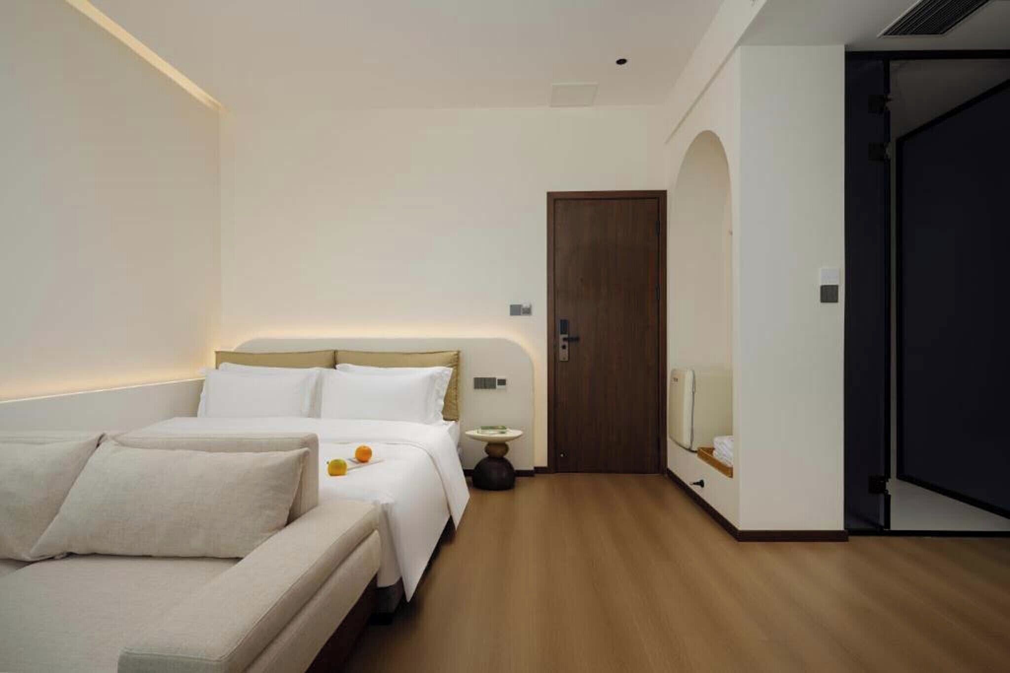 Une chambre d’hôtel moderne, équipée d’un lit, d’un canapé, d’une petite table avec une lampe et d’une porte donnant sur une autre pièce.