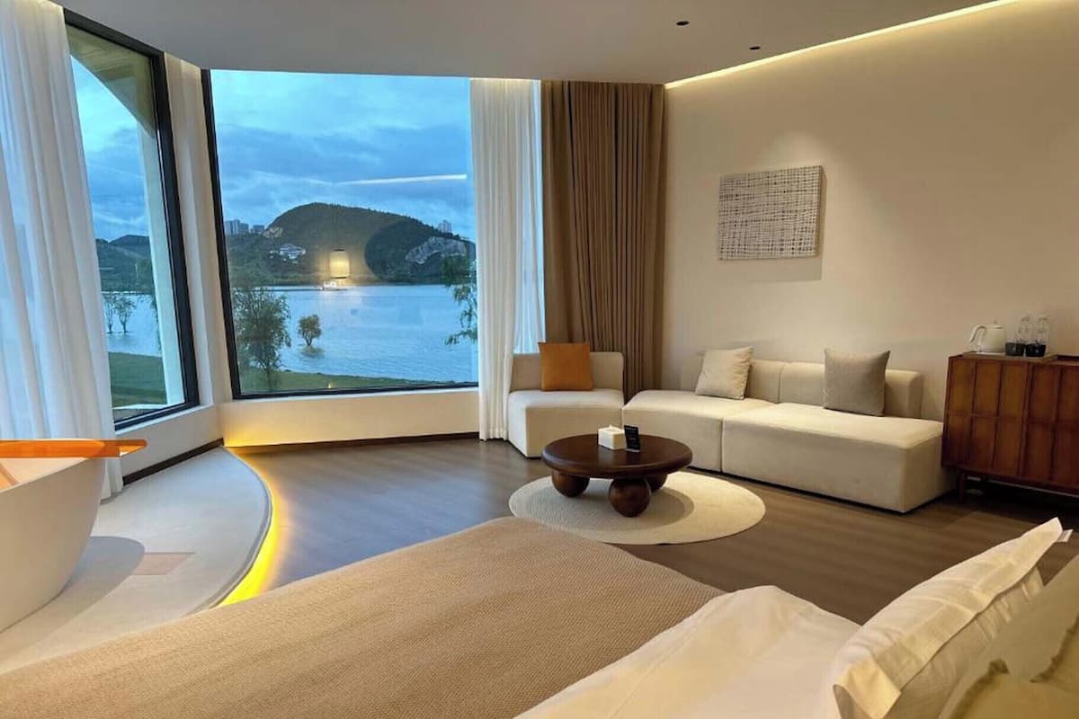 Ein modernes Hotelzimmer mit einem großen Fenster und Blick auf einen See und Hügel. Das Zimmer verfügt über ein Sofa, einen Couchtisch und ein Bett mit Kopfteil.