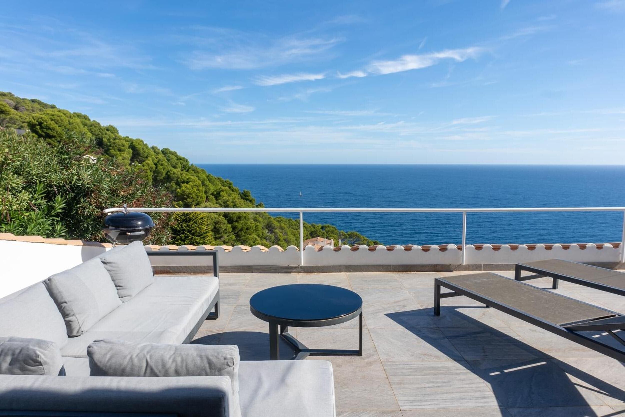 Um terraço com um sofá, uma mesa redonda e uma poltrona de descanso, com vista para uma paisagem costeira com árvores e o mar.