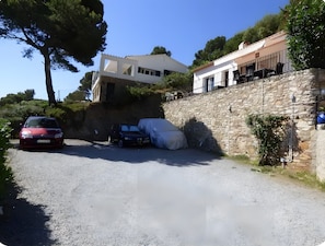 Une allée de gravier mène à un mur de pierres, avec des voitures garées et un bâtiment doté d’un toit en pente.