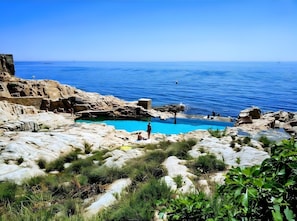 Une zone côtière rocheuse avec une piscine aux eaux bleues et limpides, entourée d’une végétation naturelle, et offrant une vue sur la mer.