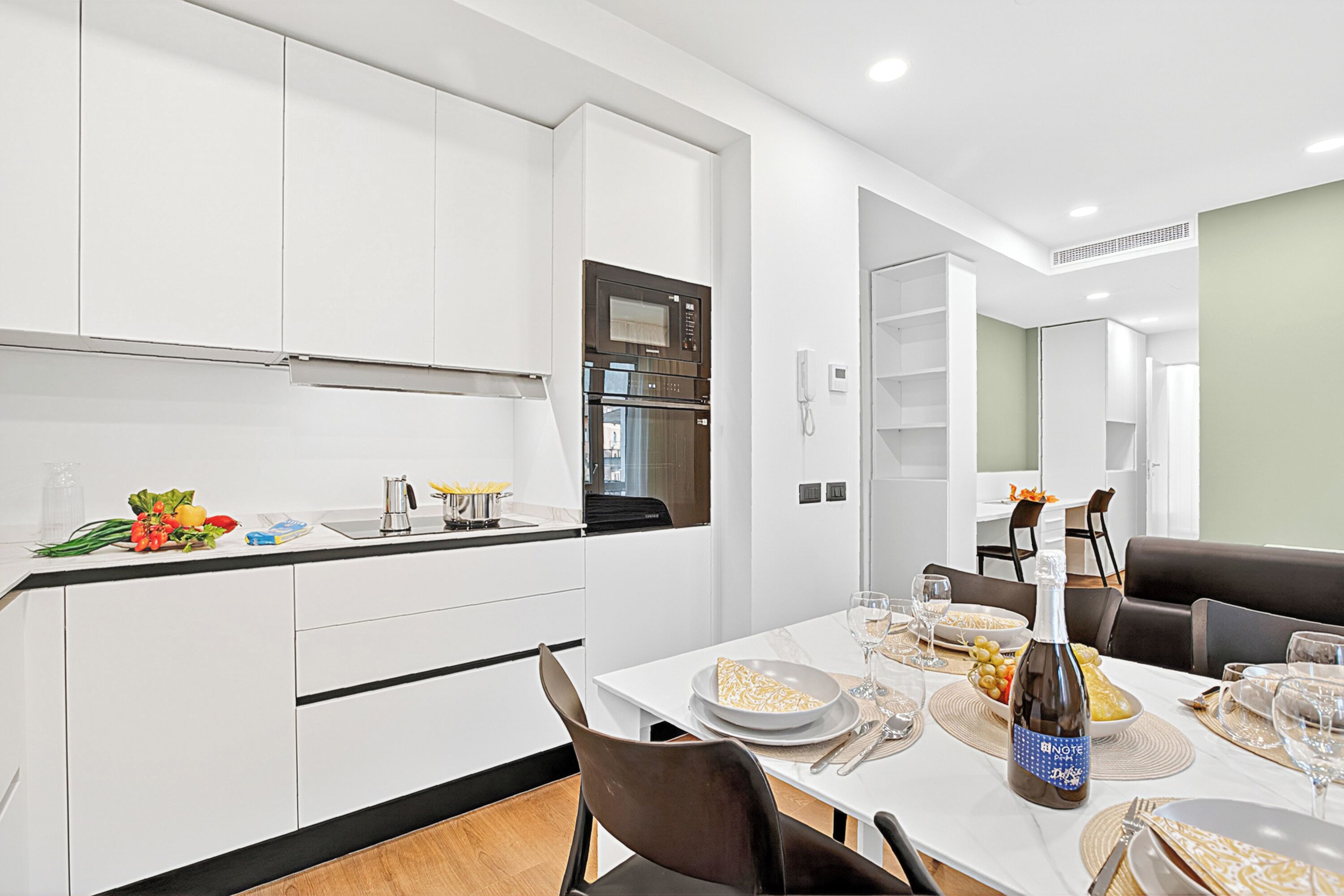 A modern kitchen with white cabinets, a built-in oven, and a dining area with a white table set for a meal.