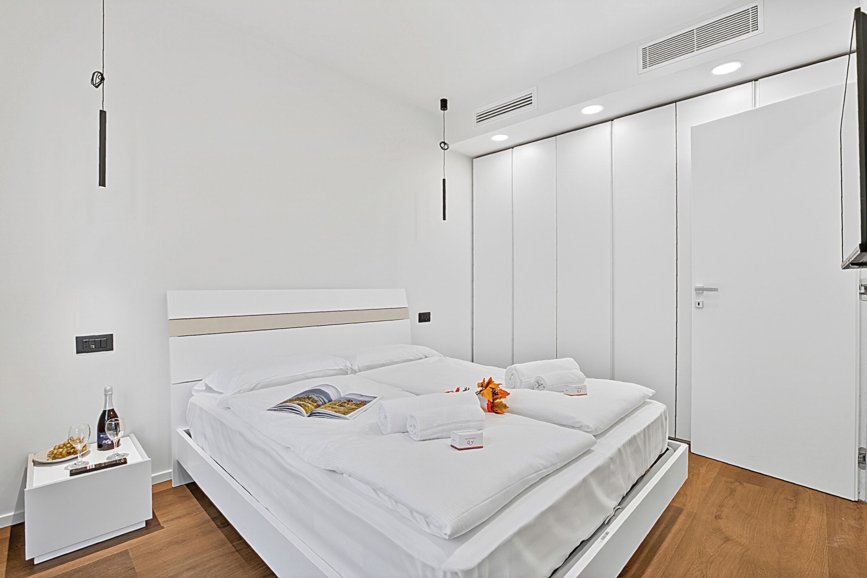 A modern hotel room with a large bed, a bedside table with a bottle and glasses, and a nightstand.