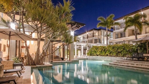 Un área de piscina con sillones y sombrillas, rodeada de palmeras y un edificio con balcones.