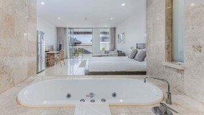Un baño moderno con una bañera grande, cabina de ducha de vidrio y vistas a un dormitorio con cama y balcón.