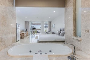 A modern bathroom with a large bathtub, a glass shower enclosure, and a view of a bedroom with a bed and a balcony. - Little Palms at Santai - 309 (Casuarina)