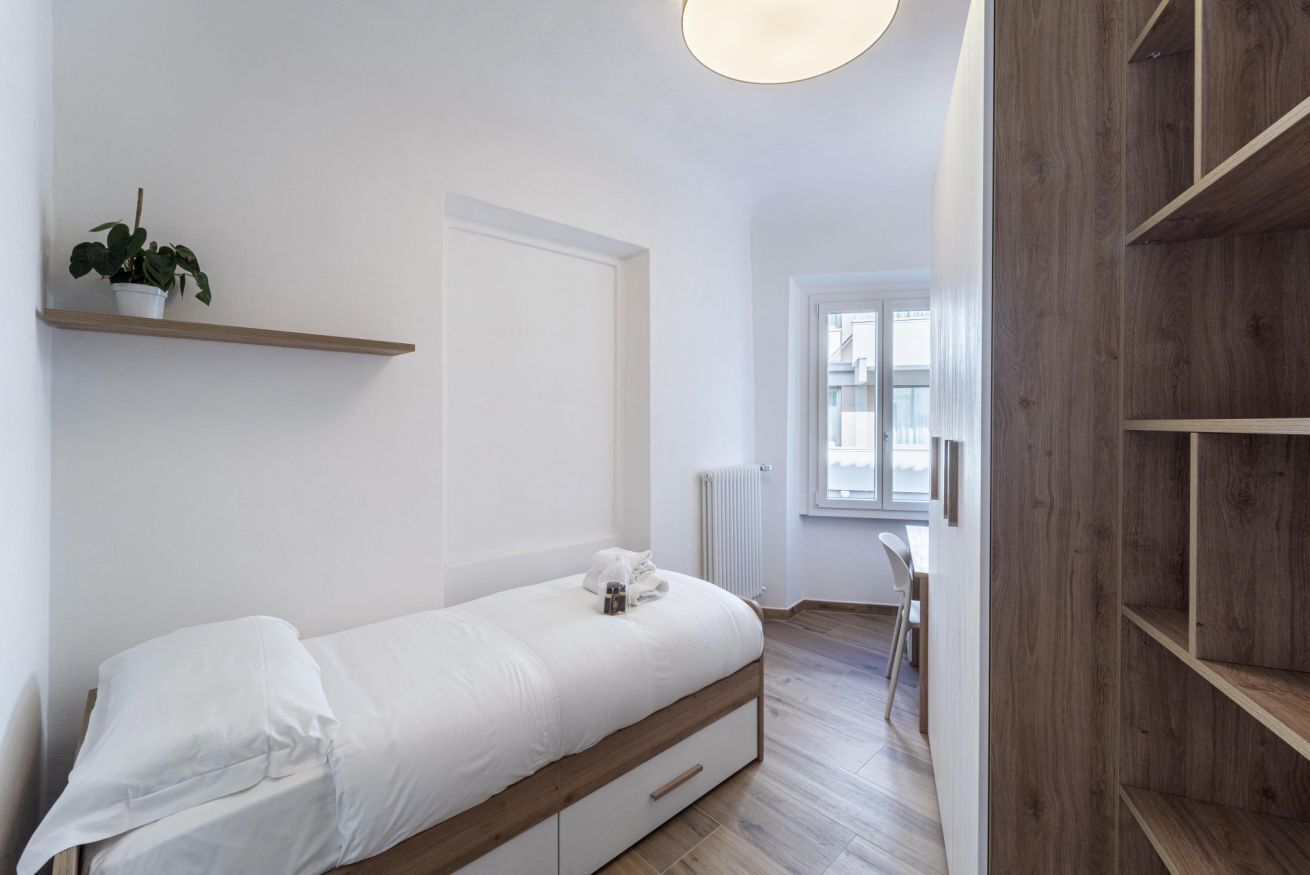 A minimalist bedroom with a single bed, a small bedside table, a radiator, and a wooden shelf.