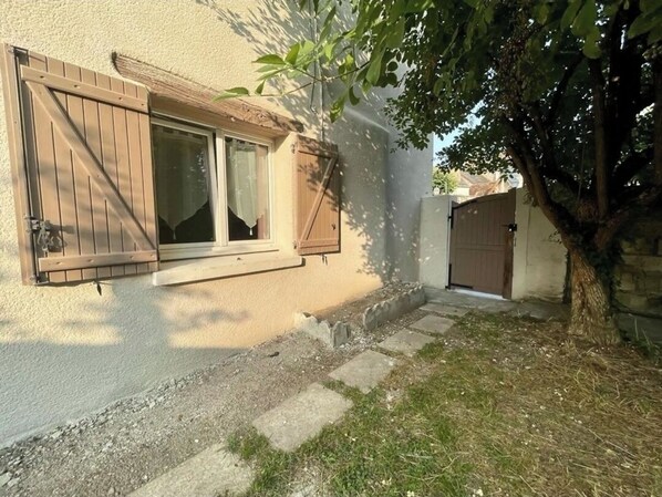 Une maison avec un volet en bois, un chemin en béton et un arbre dans la cour.