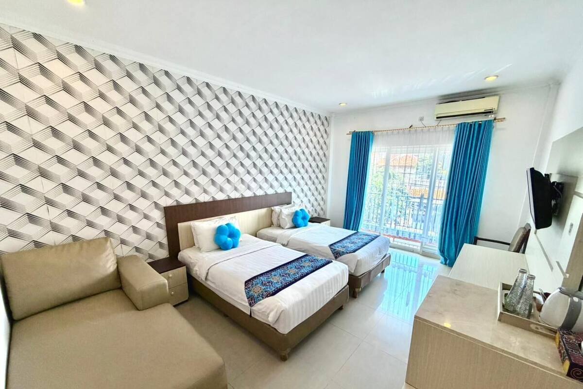 A hotel room with a patterned wall, two beds with blue pillows, a sofa, a desk, and a TV.