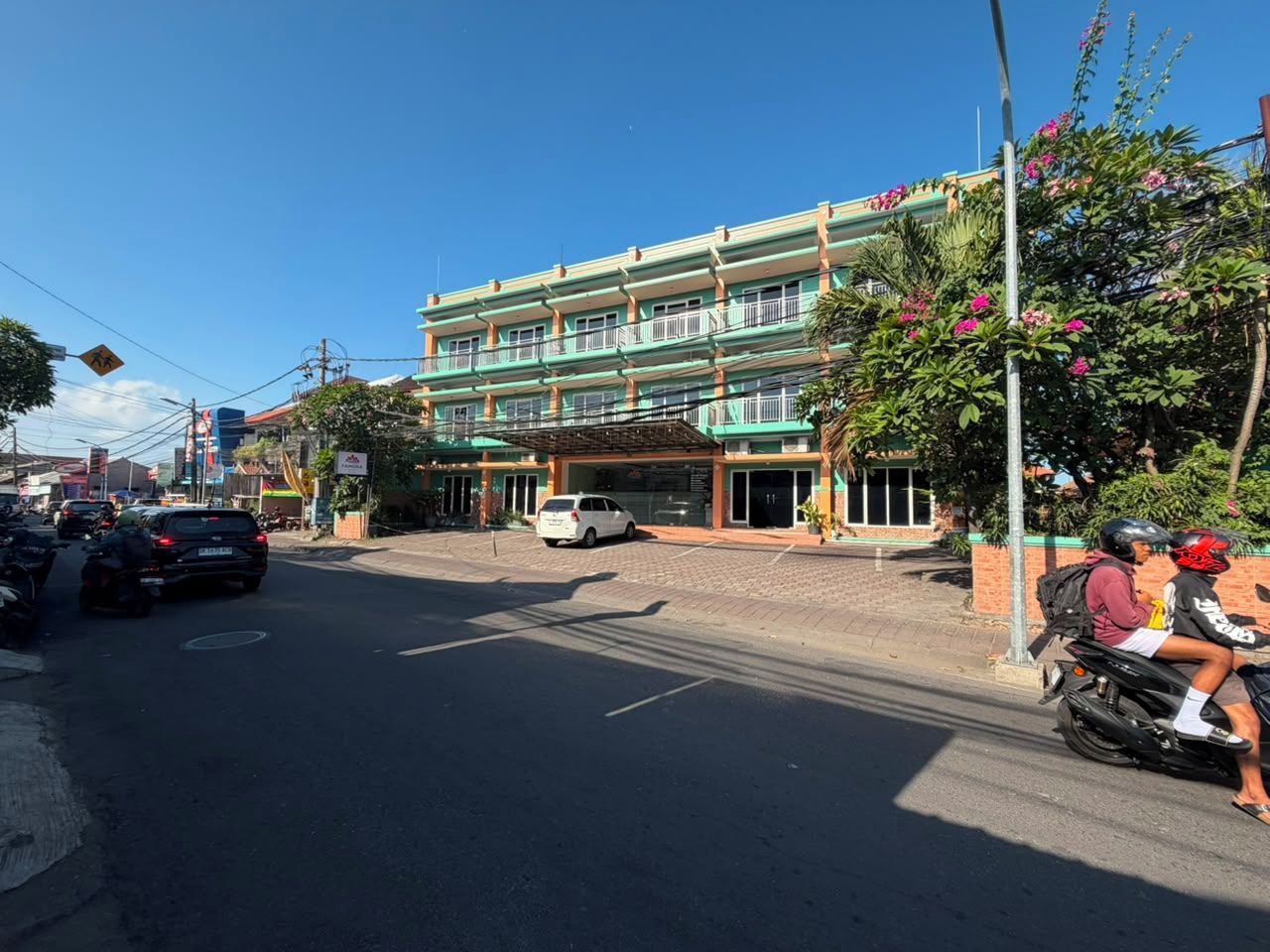 Un edificio de varios pisos con fachada verde, coches estacionados y una motocicleta con dos ocupantes.