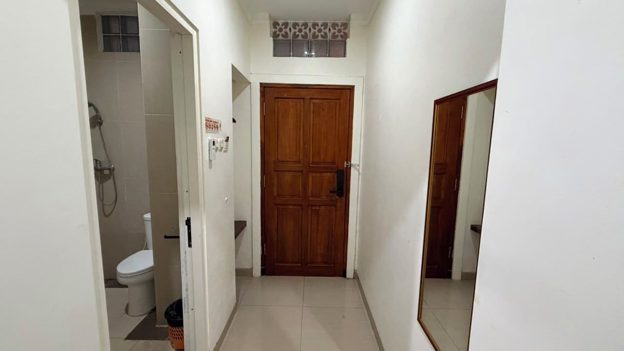 A narrow hallway with a wooden door at the end, a white toilet with a shower in the adjacent room, and a trash bin.
