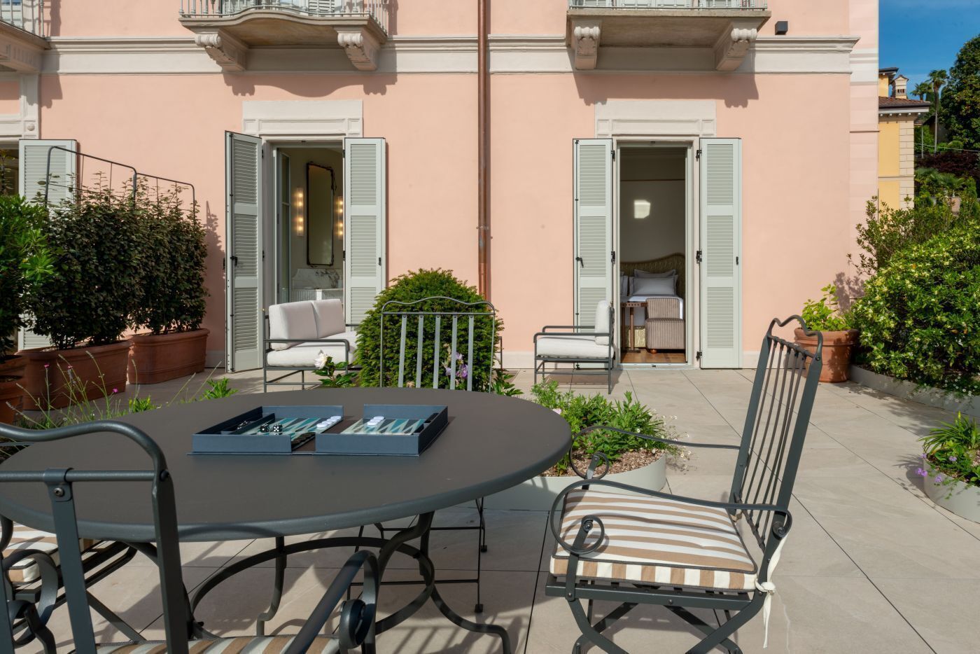 Un patio con una mesa redonda y sillas, un pequeño jardín y un edificio con contraventanas abiertas.