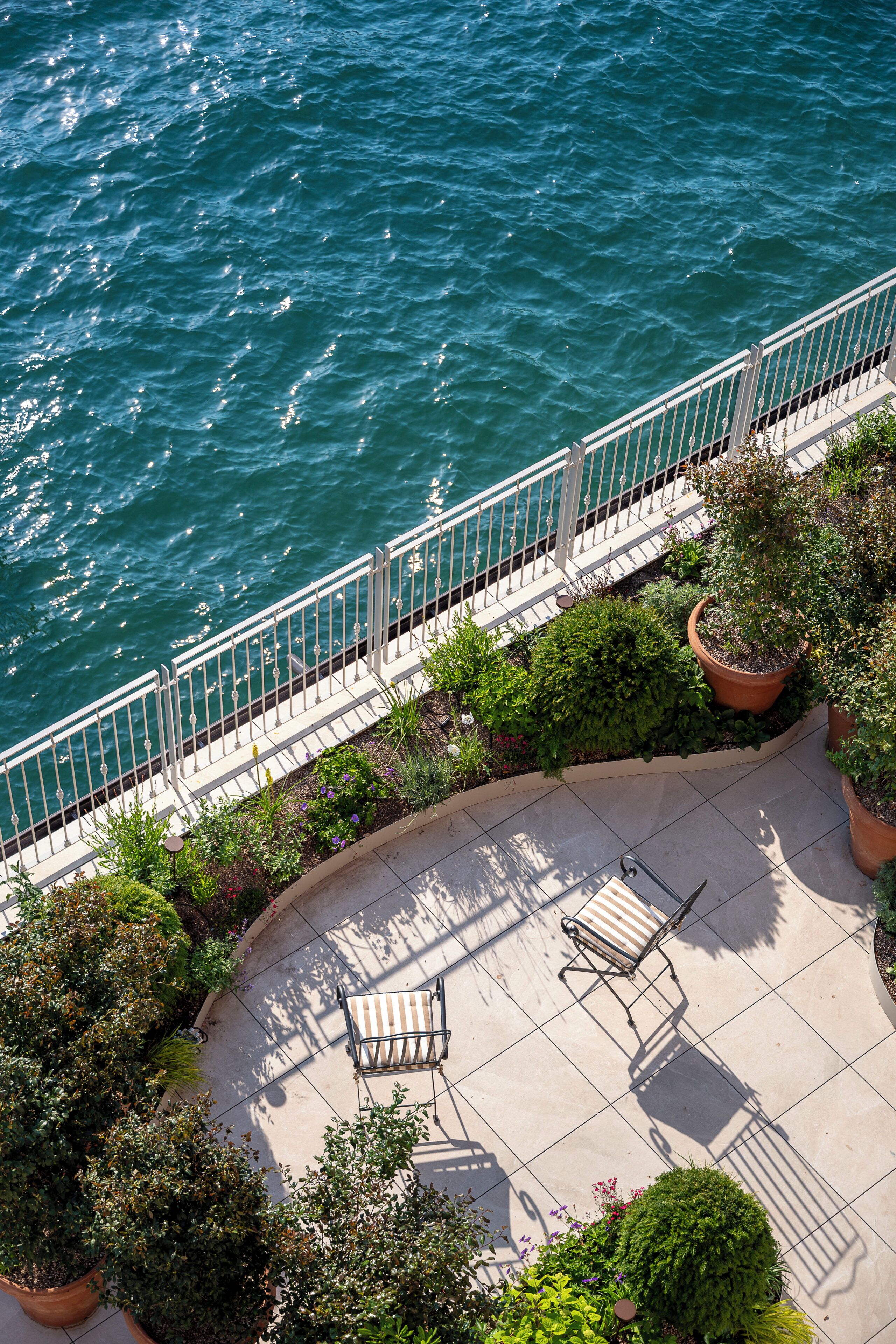 A patio with a railing overlooking a body of water, potted plants, and two chairs.