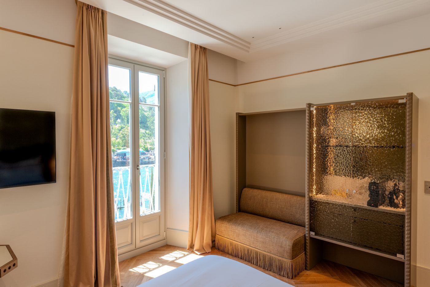 Un dormitorio con cama, televisor, banco, pared decorativa y ventana con cortinas.