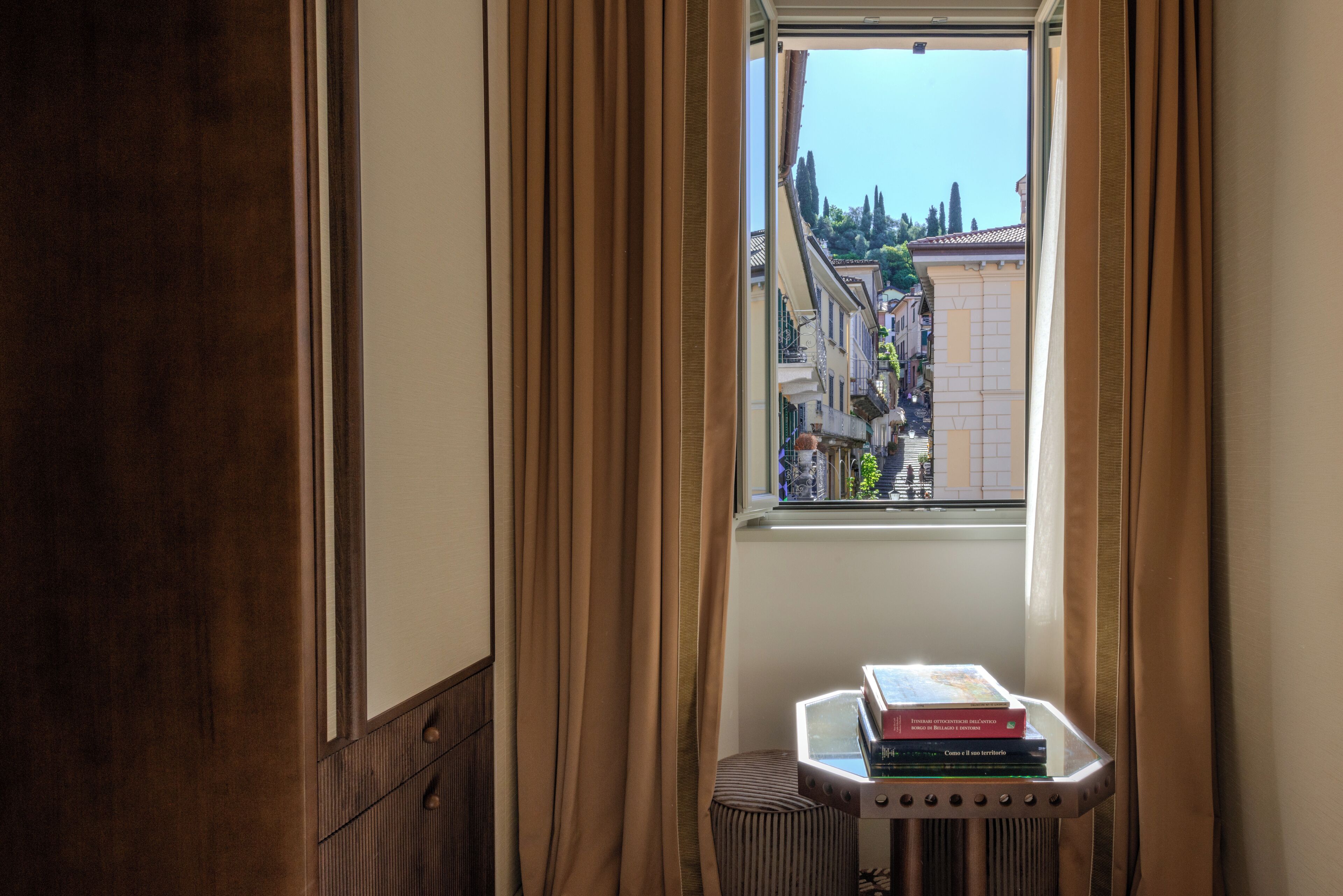 Una habitación con ventana, cortinas, un radiador y una pila de libros sobre una mesita.