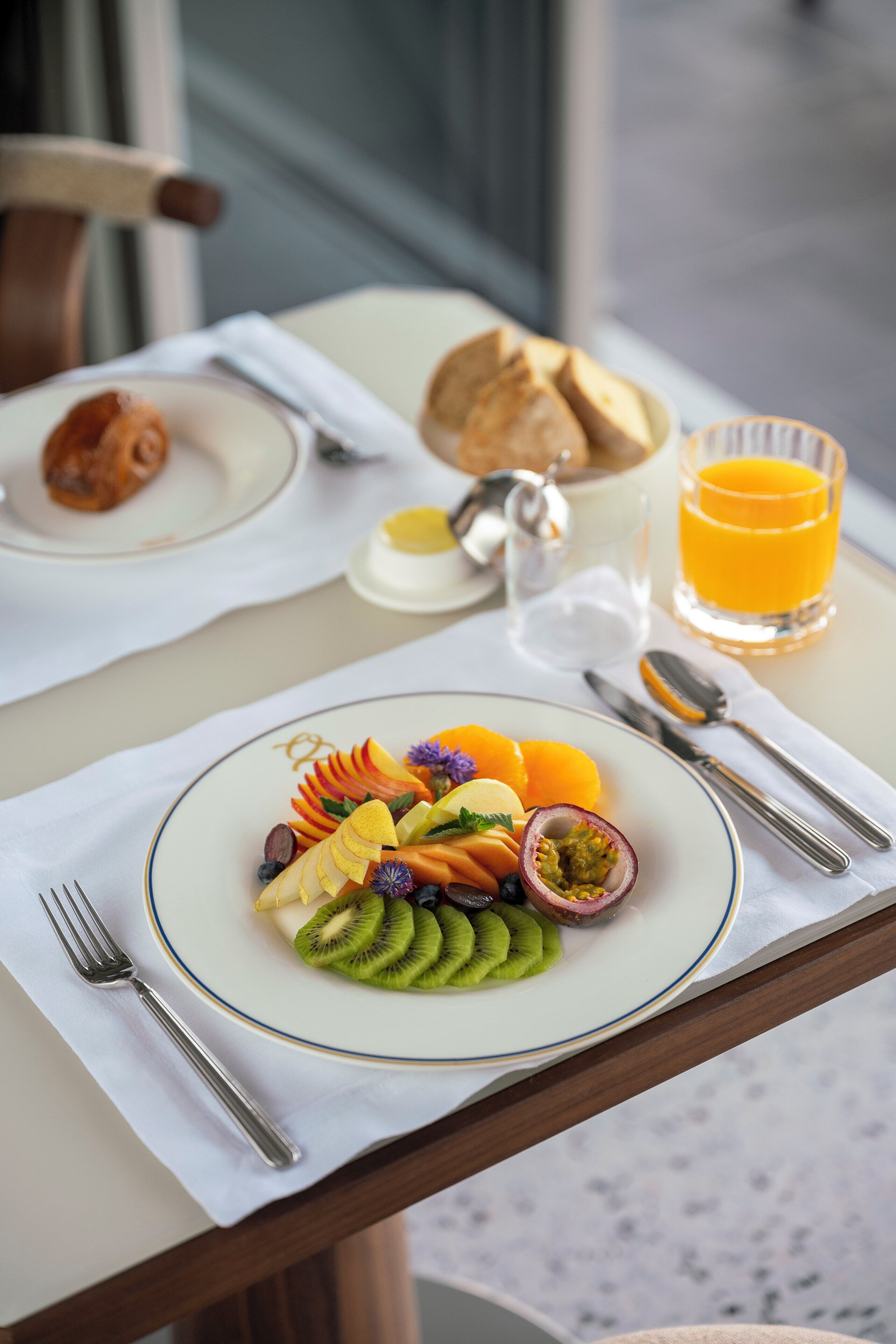 Ein Frühstückstisch mit einer Fruchtschale, einem Glas Orangensaft und einem Brötchenkorb.