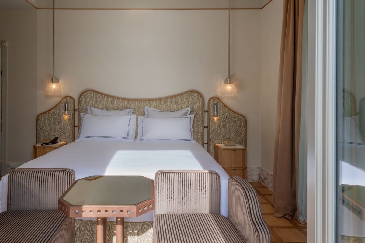 A hotel room with a large bed, two bedside tables, a small table with chairs, and a large window.