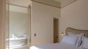 A bedroom with a bed, a nightstand, a wall-mounted lamp, and a bathroom with a bathtub and shower. - Palazzo Genazzini (Bellagio)