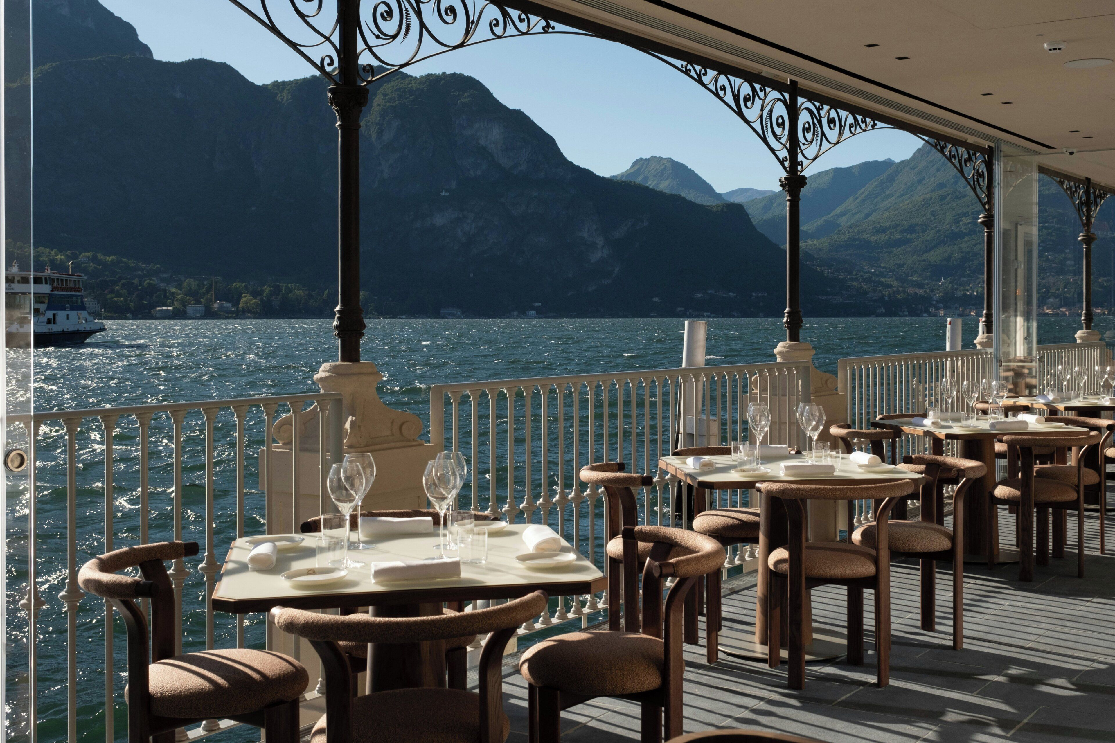 Un comedor con mesas y sillas, una barandilla y vista a un lago con montañas al fondo.