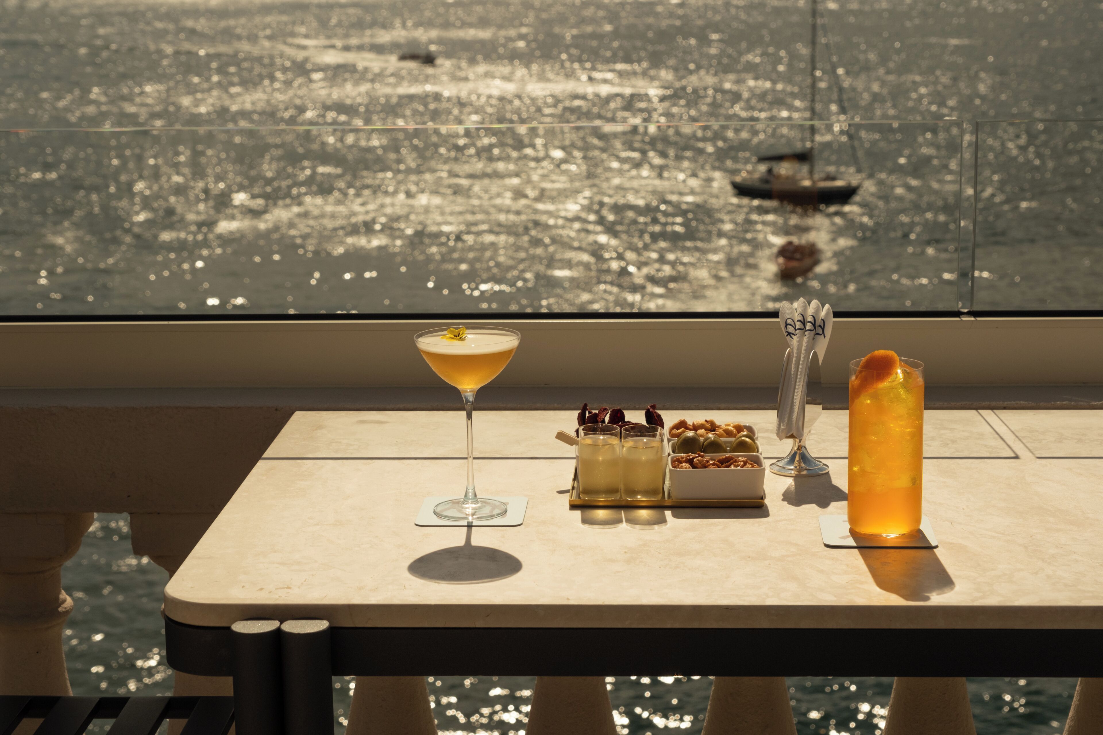 Een tafel met een martini, een hoog glas met een drankje, en een schaal met desserts en een karaf, alles op een balkon met uitzicht op het water.