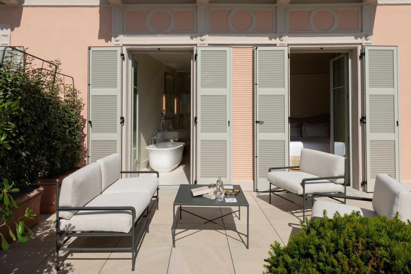 Une terrasse extérieure avec un canapé, des fauteuils et une table, une baignoire et une chambre à coucher visible à travers des portes ouvertes.