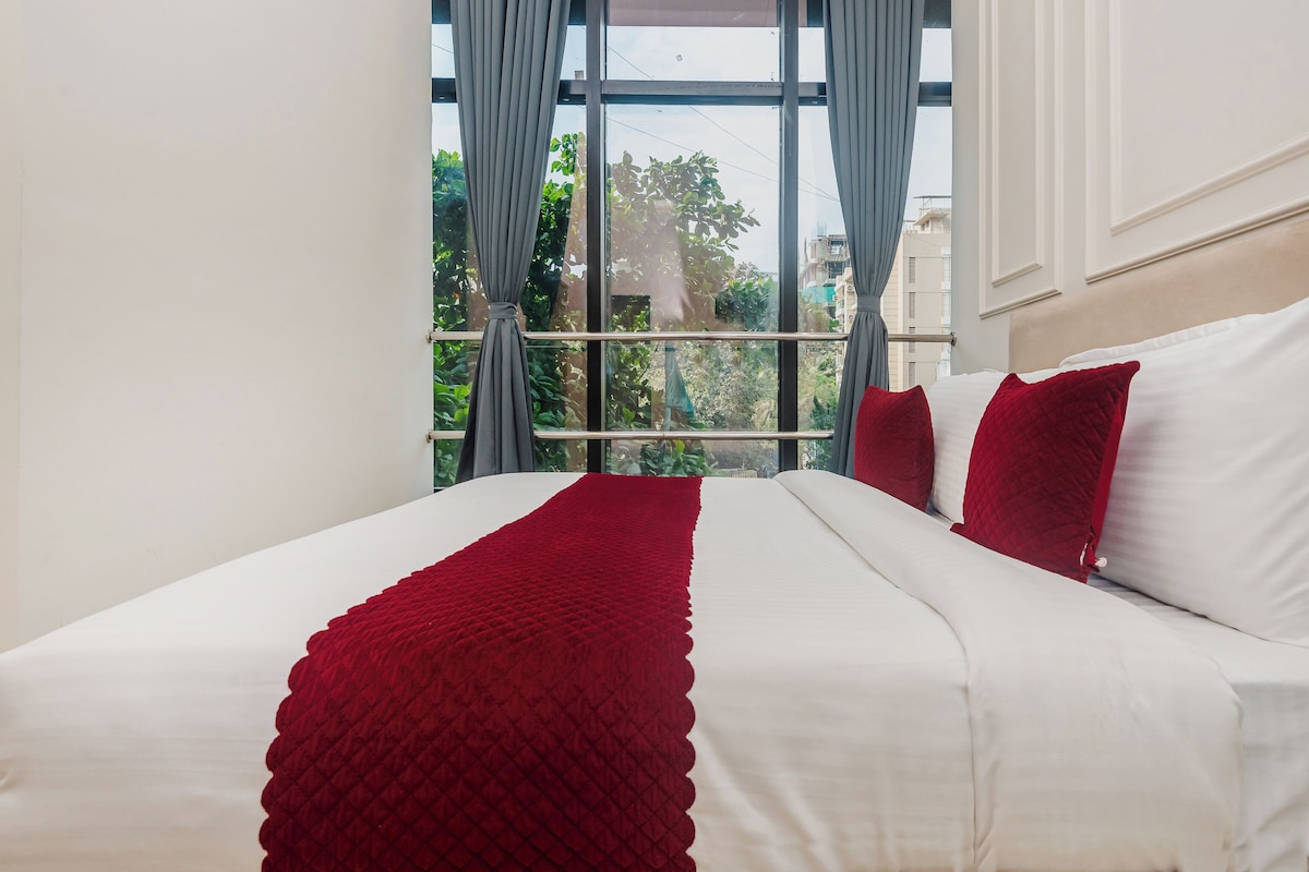 Hotelkamer met een groot bed, wit bedlinnen, rode kussens en uitzicht door een raam met gordijnen.