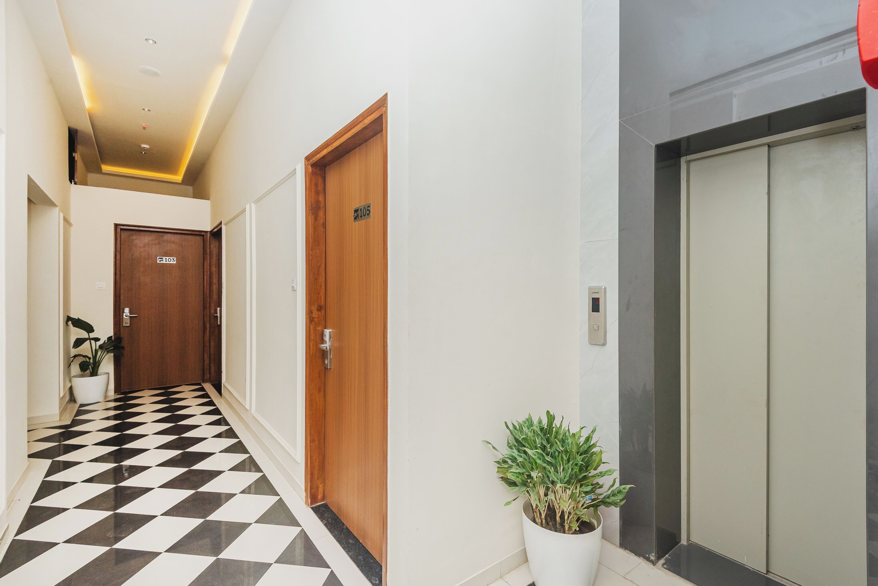 A hallway with a checkered floor, wooden doors, a potted plant, and a modern elevator.
