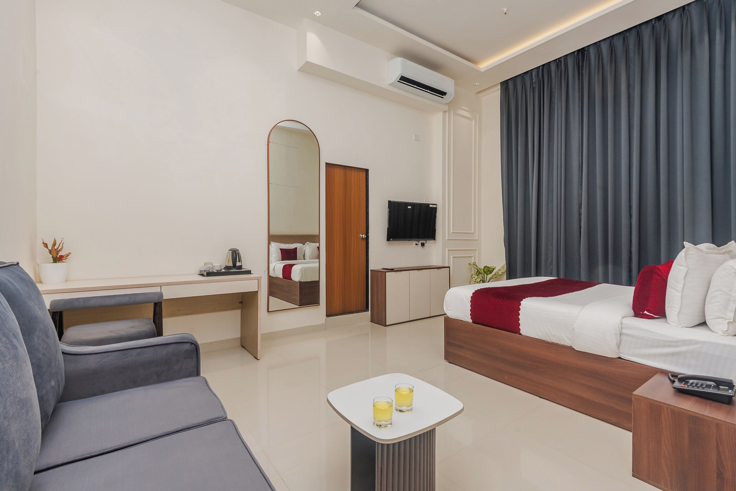 Una camera d'albergo moderna con un divano, un tavolino con bicchieri, un letto con copriletto rosso, una TV e uno specchio.