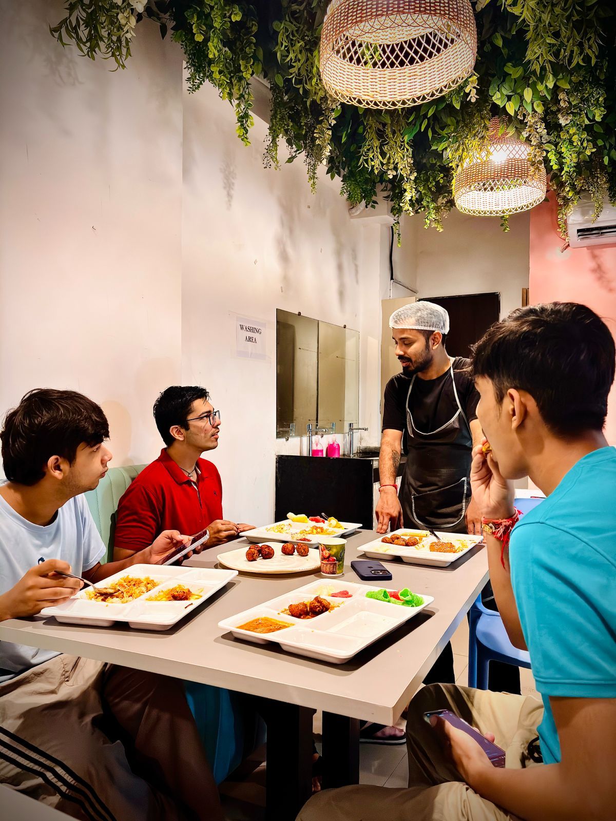 A group of people dining at a table with various dishes, a person standing possibly serving, and a sign indicating a washing area.