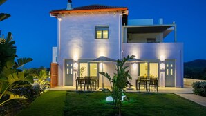 A modern two-story house with a flat roof, large windows, and a well-lit patio area with outdoor furniture. - Sweet Sissi 2BR Residence with Jacuzzi Sleeps 6 (Sisi)