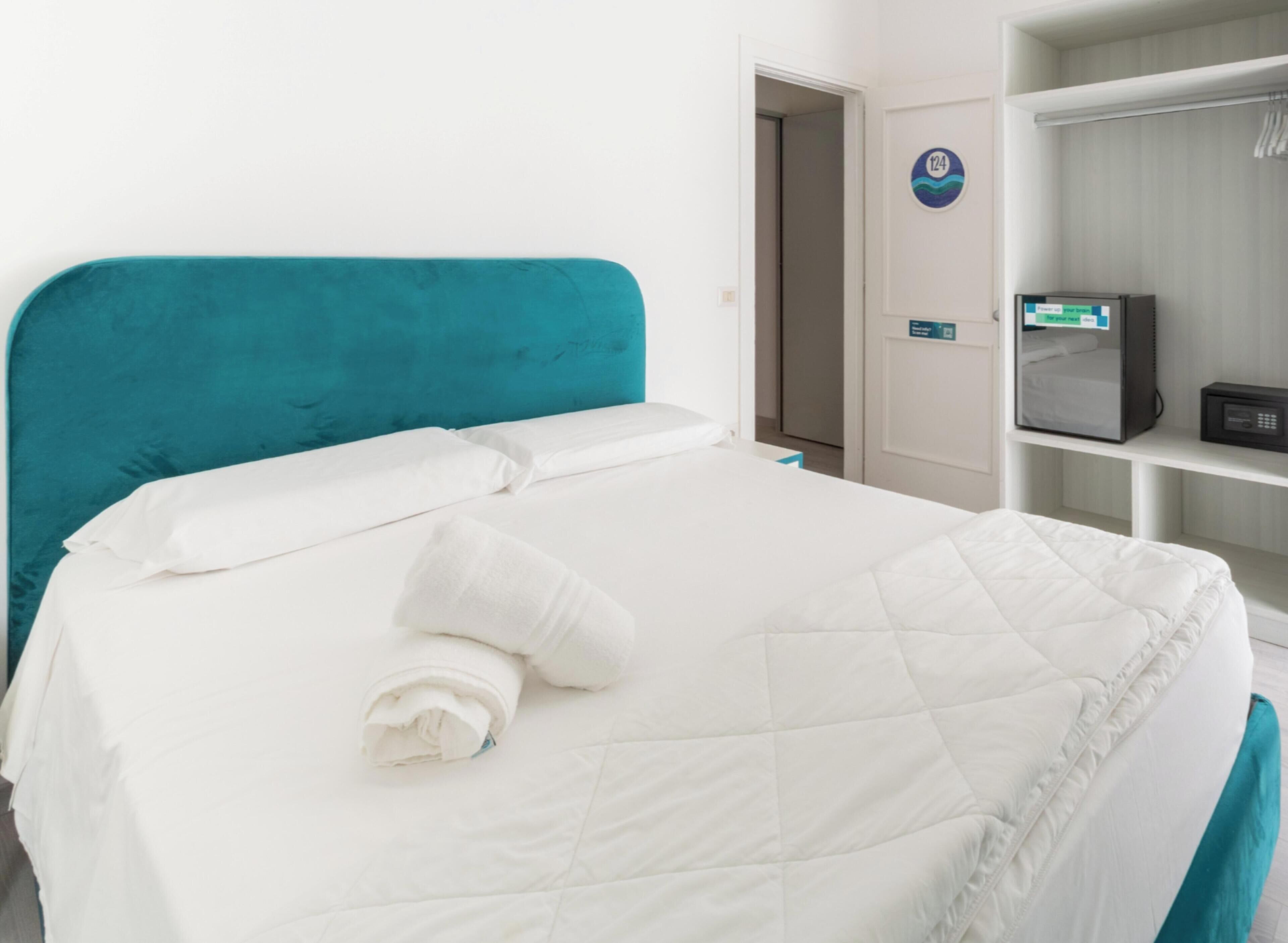 A neatly made bed with white bedding and a teal headboard, a small bedside table with a remote control, and a door with a sign indicating room number 124.