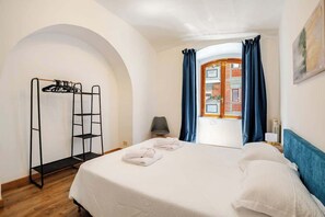 Camera d'albergo con un letto, un armadio in metallo nero, una sedia e una finestra con tende azzurre.
