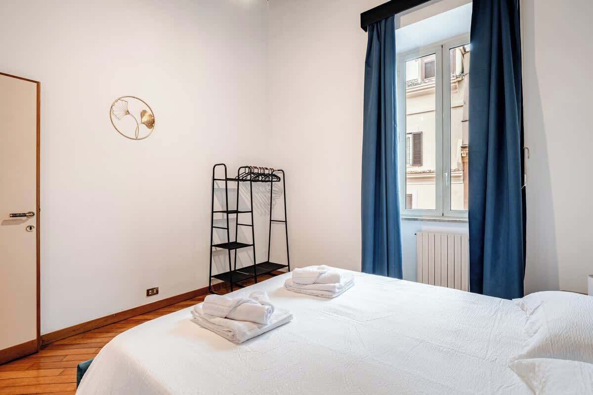 Ein Hotelzimmer mit einem Bett, zwei Handtüchern, einem Metallregal und einem Fenster mit blauen Vorhängen.