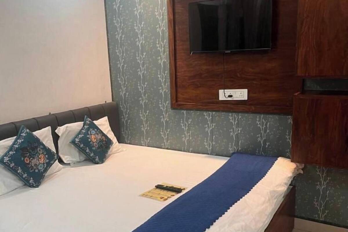 Una cama bien hecha con una manta azul y almohadillas florales, un televisor montado en un panel de madera encima, y un armario de madera a la derecha.