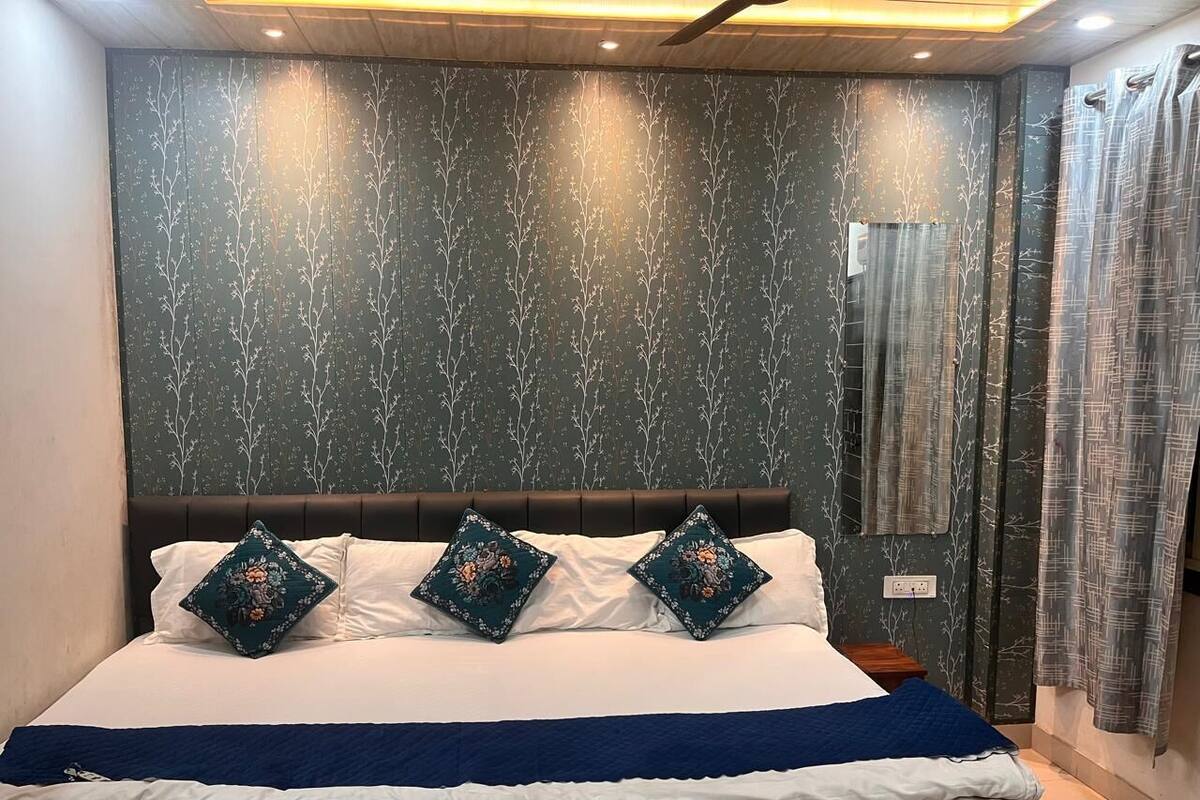 Una cama bien tendida con sábanas blancas y dos almohadones decorativos, cabecera oscura y pared con estampado de árbol.
