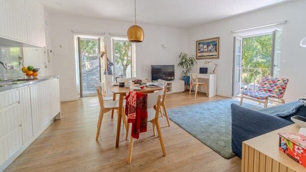 Eine moderne Küche mit weißen Schränken, ein Essbereich mit Holzsesseln, ein Fernseher und ein großes Fenster.