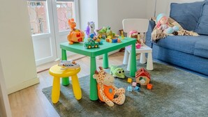 Ein Spielbereich für Kinder mit einem grünen Tisch, einem gelben Hocker und verstreuten Spielsachen auf dem Boden.