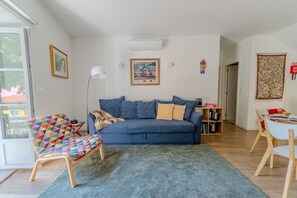 Ein Wohnzimmer mit blauem Sofa, einem bunten Sessel, einem kleinen Tisch und einem Bücherregal.
