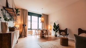 A cozy living room with a wooden floor, a dining table with chairs, a sofa, a large window with curtains, and a hanging light fixture. - Luga Homes - Augustusplatz (Leipzig)