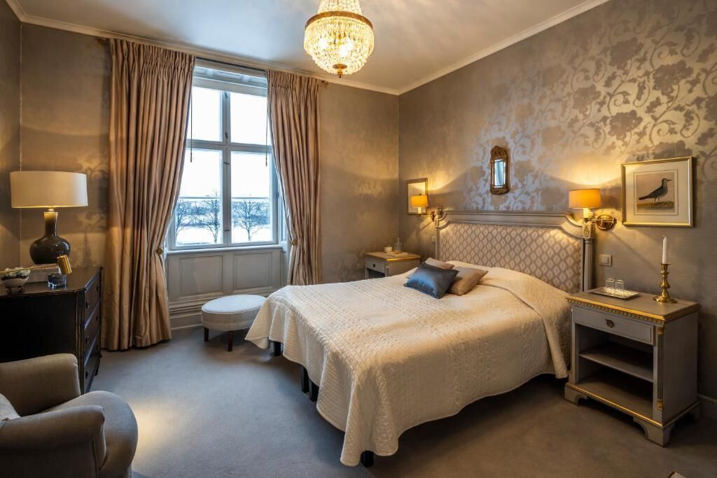 A hotel room with a large bed, a chandelier, a window with curtains, a nightstand with a lamp, and a framed picture on the wall.