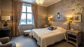 A hotel room with a large bed, a chandelier, a window with curtains, a nightstand with a lamp, and a framed picture on the wall.