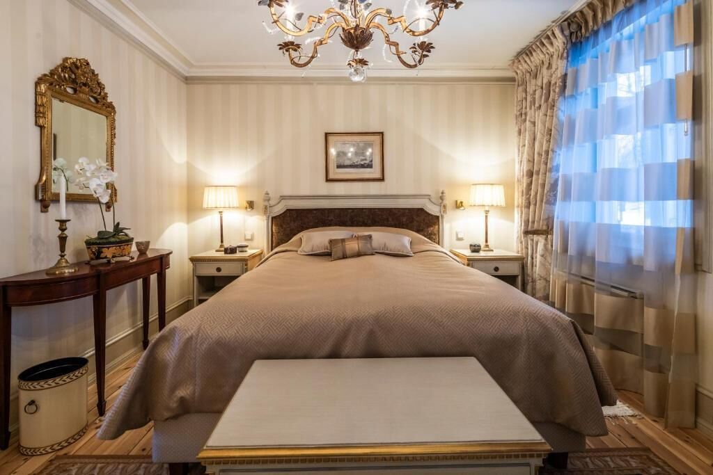 Habitación de hotel con una cama grande, dos mesitas de noche con lámparas, un candelabro, un espejo y una ventana con cortinas.